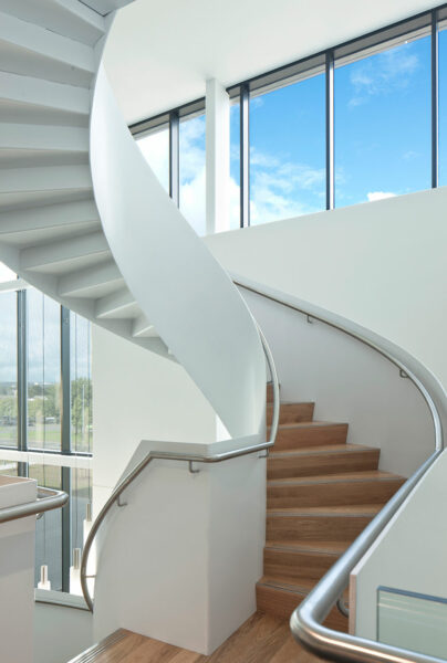 Spiral wooden staircase with white balustrade and windows above letting natural light shining on.